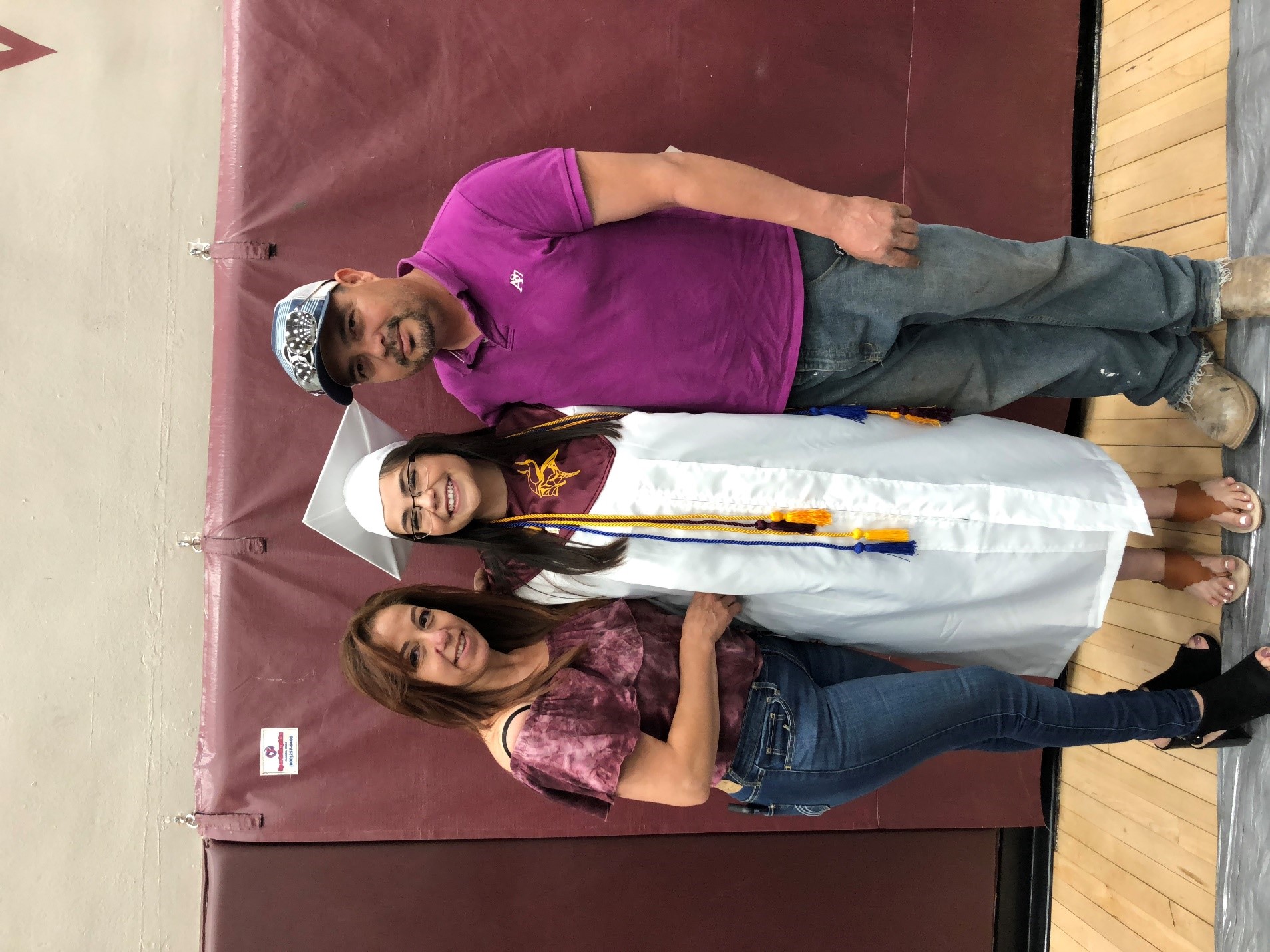 Arlin Arpero stands in a gymnasium, smiling at the camera and wearing a white cap and gown with tassels. A woman wearing a maroon shirt and jeans stands to Arlin's left. A man wearing a maroon shirt and a cap stands to Arlin's right.