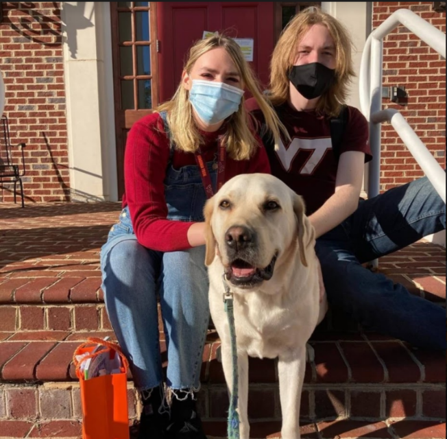 Two people, wearing surgical masks, sit on a stoop. Hannah, on the left, wears a red sweater and jeans; on the right, James wears a Virginia Tech shirt. They are petting a yellow lab that sits in front of them named Derek.