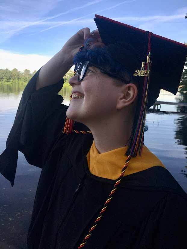 Ila White, wearing cap and gown with a 2021 tassel, smiles to the left of the image with a blue sky and lake in the background.