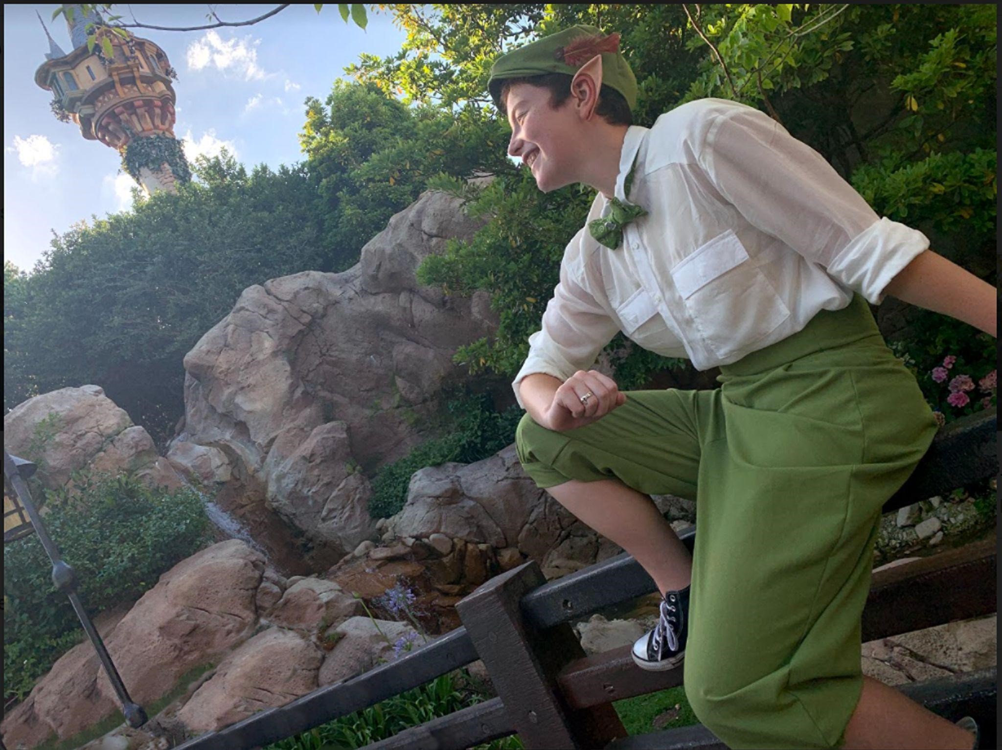 Ila White, sitting on a fence with a forest scene in the background, wearing green pants, a white shirt, a green bowtie, a green felt hat, and pointed ears.