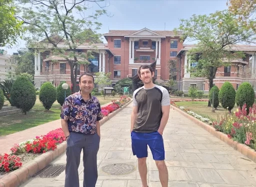 Two men stand in the foreground hands in pockets. Behind them is a large brick building with columns. Brilliant red flowers line the stone walkway to the building along with trees and well manicured bushes. 