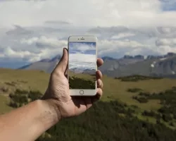 photo of a white hand holding a mobile phone in front of a landscape.