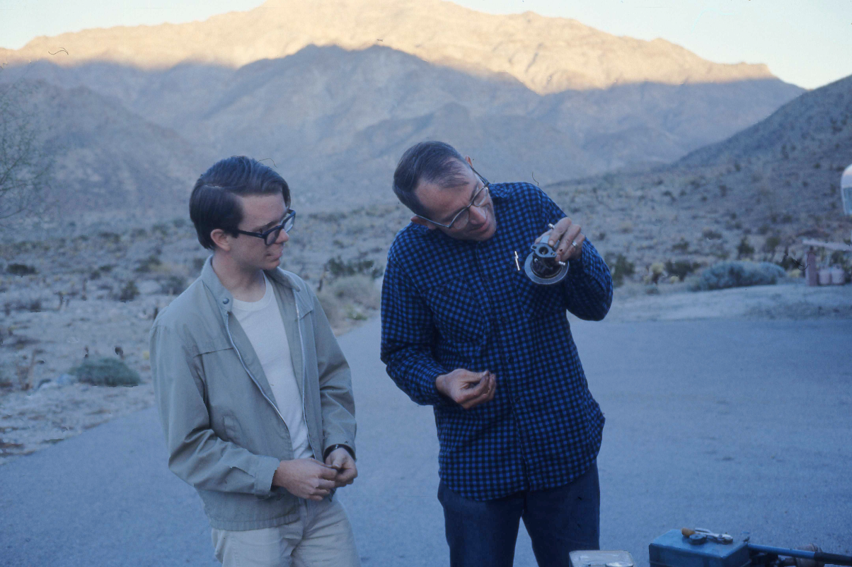 two men inspect a small piece of equipment with mountains in the background