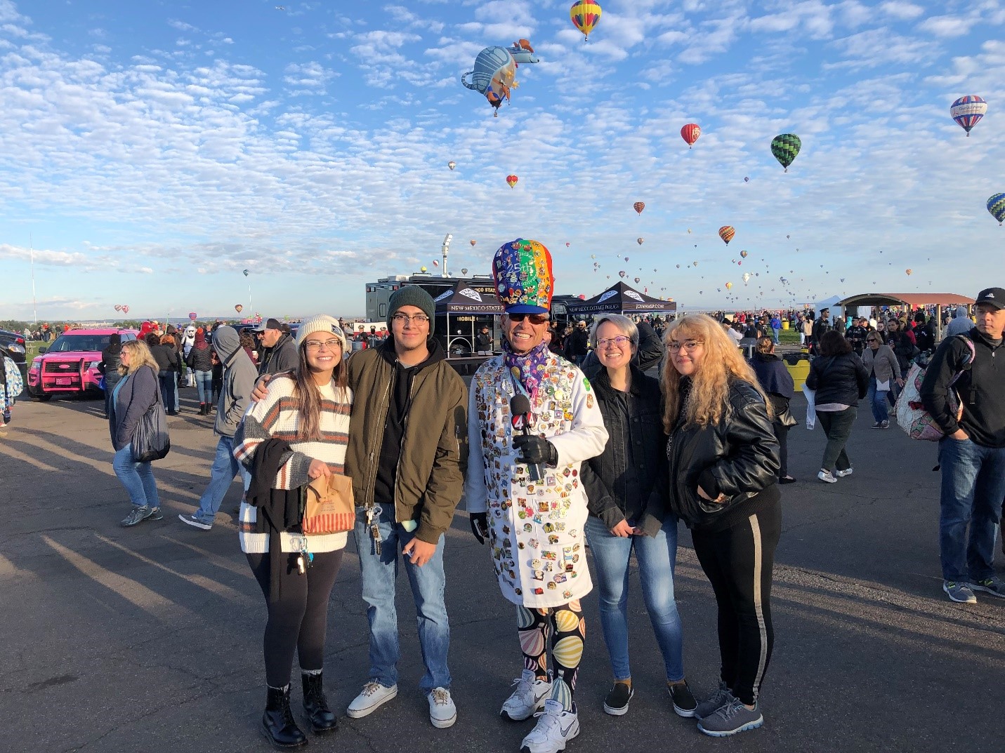 5 people stand smiling at the camera. They are outdoors with a blue sky filled with many hot air balloons. Arlin Arpero stands to the left, wearing a sweater and a hat, next to a man in a hoodie and hat. In the center, a man wears a bright rainbow cap and a white coat covered in colorful pins. Two women stand to the right in the image, wearing black jackets.