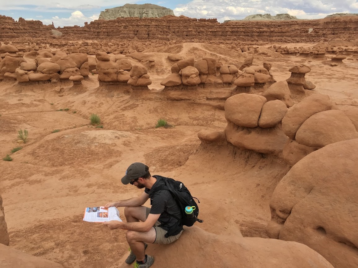 Philip, wearing a cap and hiking backpark, sits on a red sandstone rock in a valley of similar smooth red rocks and reads a pamphlet.