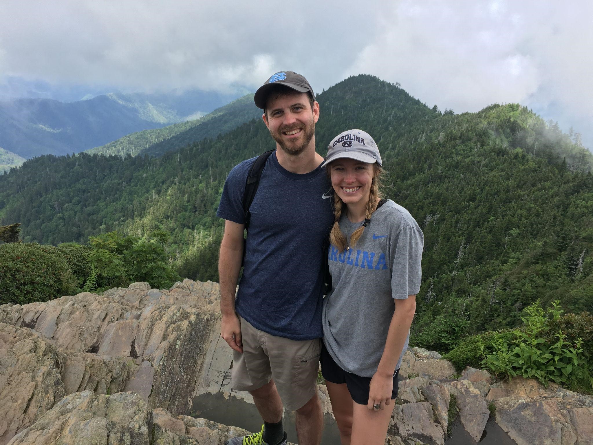 Two people, Philip and Allie, stand in front of a rocky mountaintop surrounded by wooded peaks with a cloudy sky.