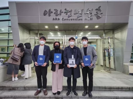 Four individuals in COVID-era masks hold certficates in front of a building with Korean lettering