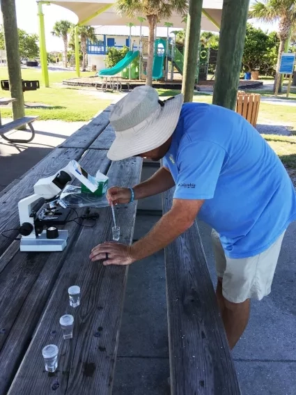 A man in a blue shirt and sun hat uses a microscope and water samples at a picnic table in a park to analyze environmental data.