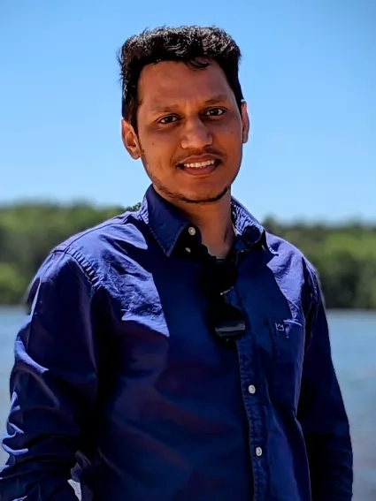 Man in blue shirt with sunglasses in collar smiles for outdoors portrait in front of water 