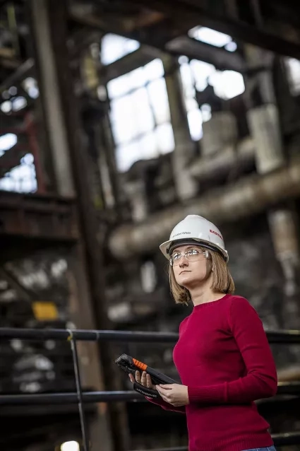 Woman in red shirt wears a hard hat and holds technological device in an industrial setting