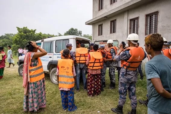 Participants in orange life vests and white helmets conduct a flood anticipatory simulation near a building and white vehicle in the Karnali Basin, Nepal.