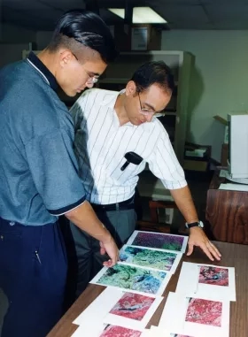 Two men standing over a table looking at prints of satellite imagery