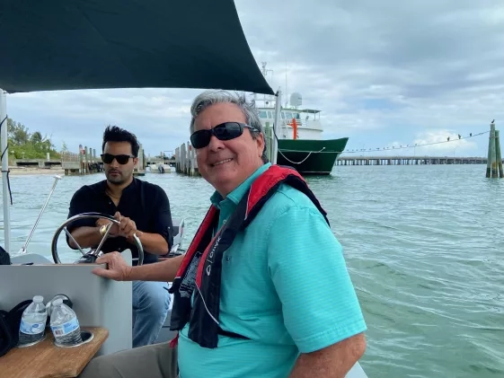 Ved Chirayath and Maury Estes are on a small electric boat in the ocean with a larger boat and dock visible behind them. They are both smiling and wearing sunglasses.