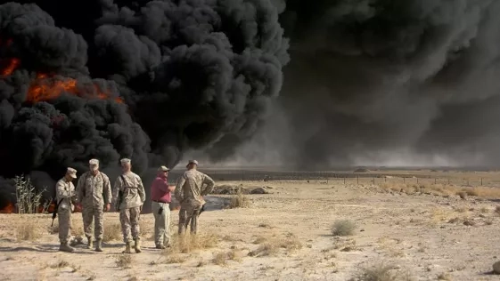 five men, four of whom are in U.S. Marine uniforms, stand in conversation near a burn pit billowing black smoke into the sky