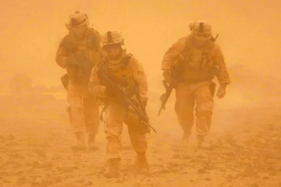 three soldiers in combat gear walk through the desert, with the image obscured by orange haze from a sand storm