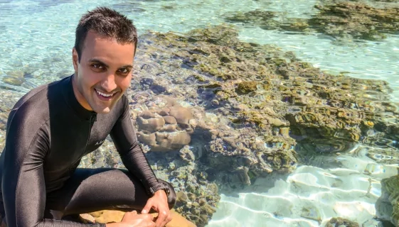 Ved is wearing a wetsuit and smiling. Behind him coral and sand are visible beneath clear rippling water.