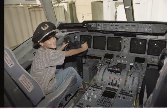 A young Ved grins as he sits in the cabin of an air or spacecraft, wearing a captains hat and pretending to steer. 