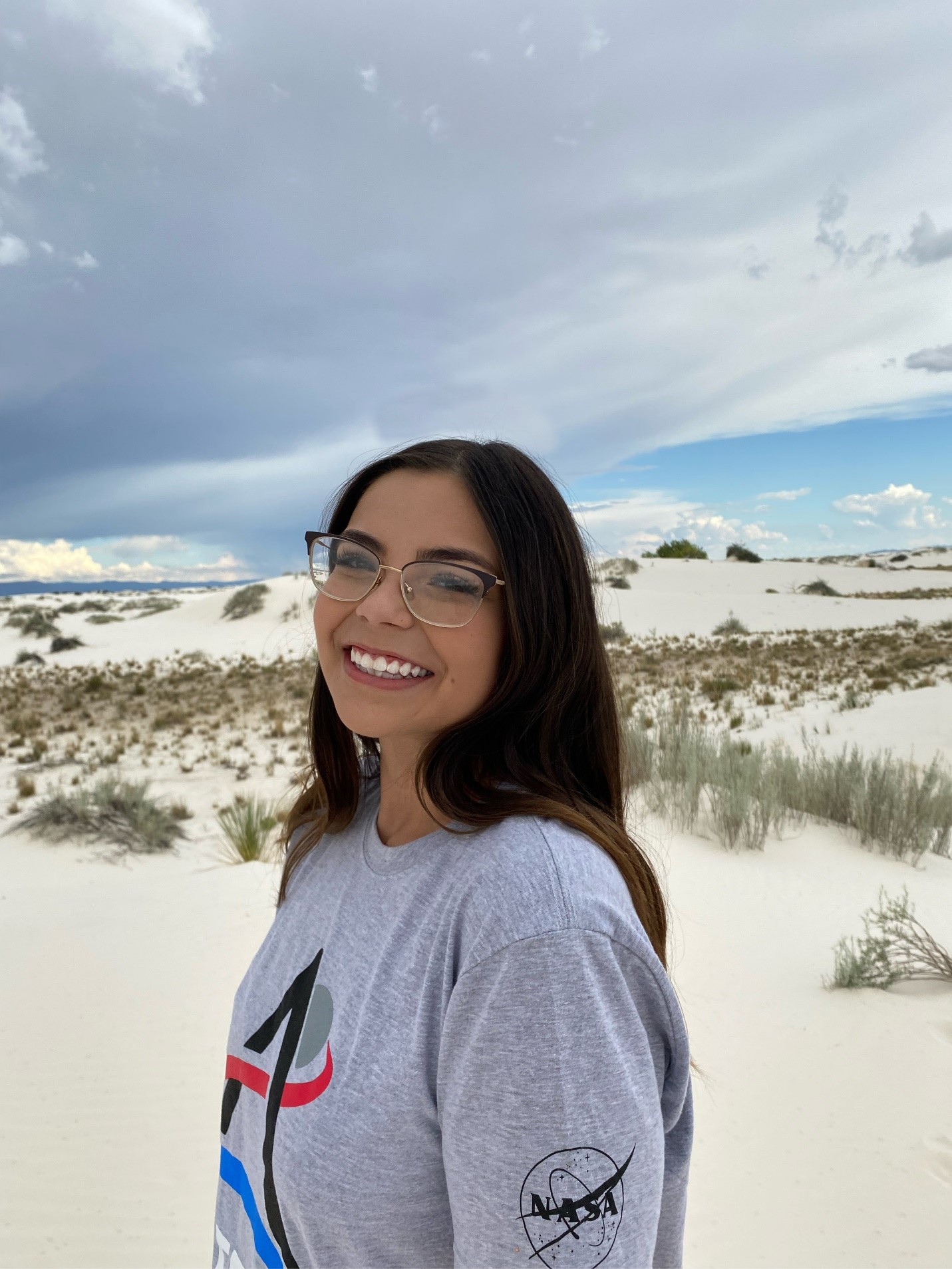 Arlin Arpero stands in a sandy desert with a cloudy sky. Arlin  looks at the camera while facing the left, and wears glasses and a grey NASA Artemis t-shirt.