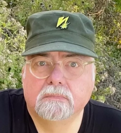 Photo of white man with a grey goatee wearing glasses and a green hat