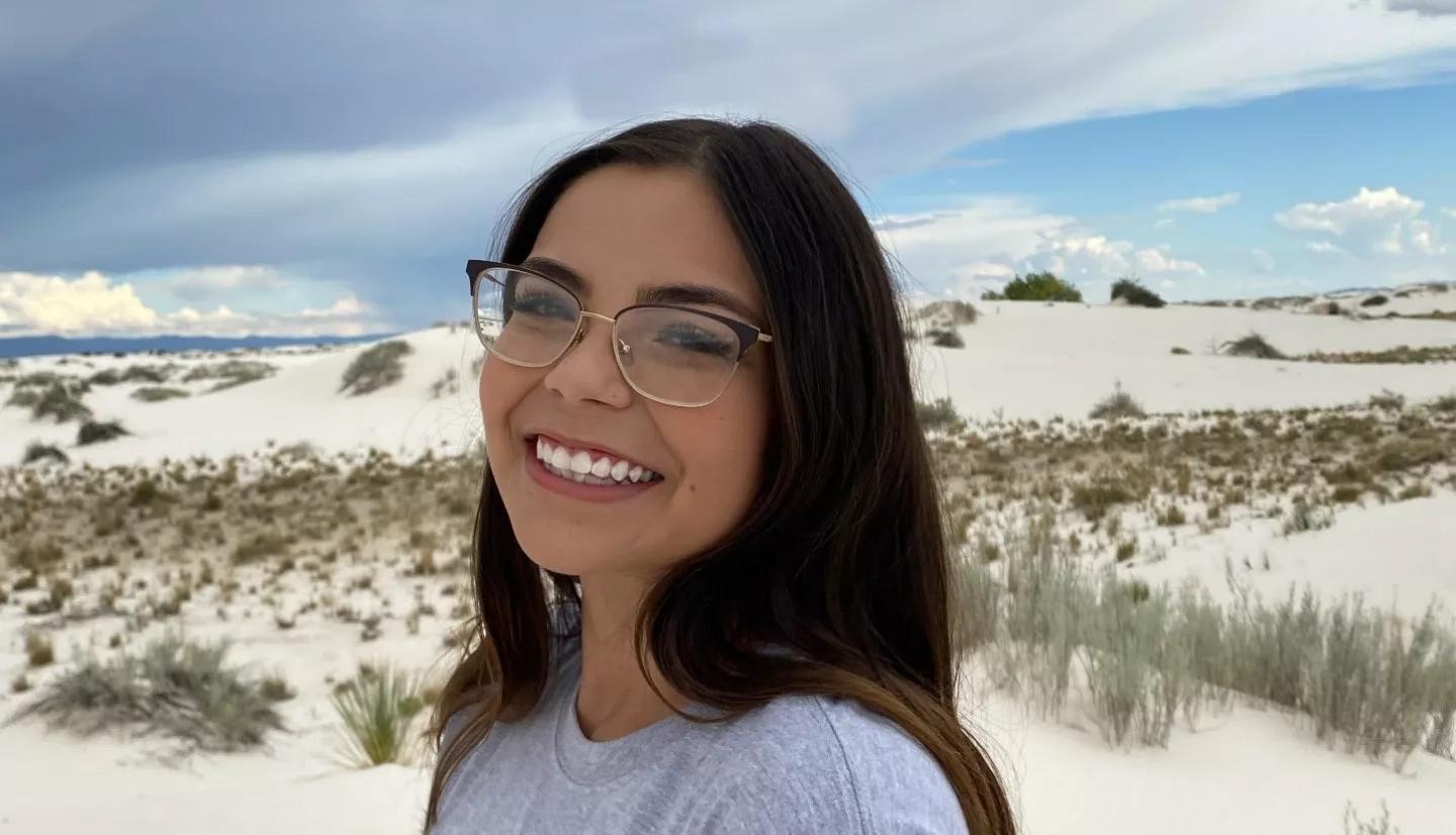 Arlin Arpero stands in a sandy desert with a cloudy sky. Arlin  looks at the camera while facing the left, and wears glasses and a grey NASA Artemis t-shirt.