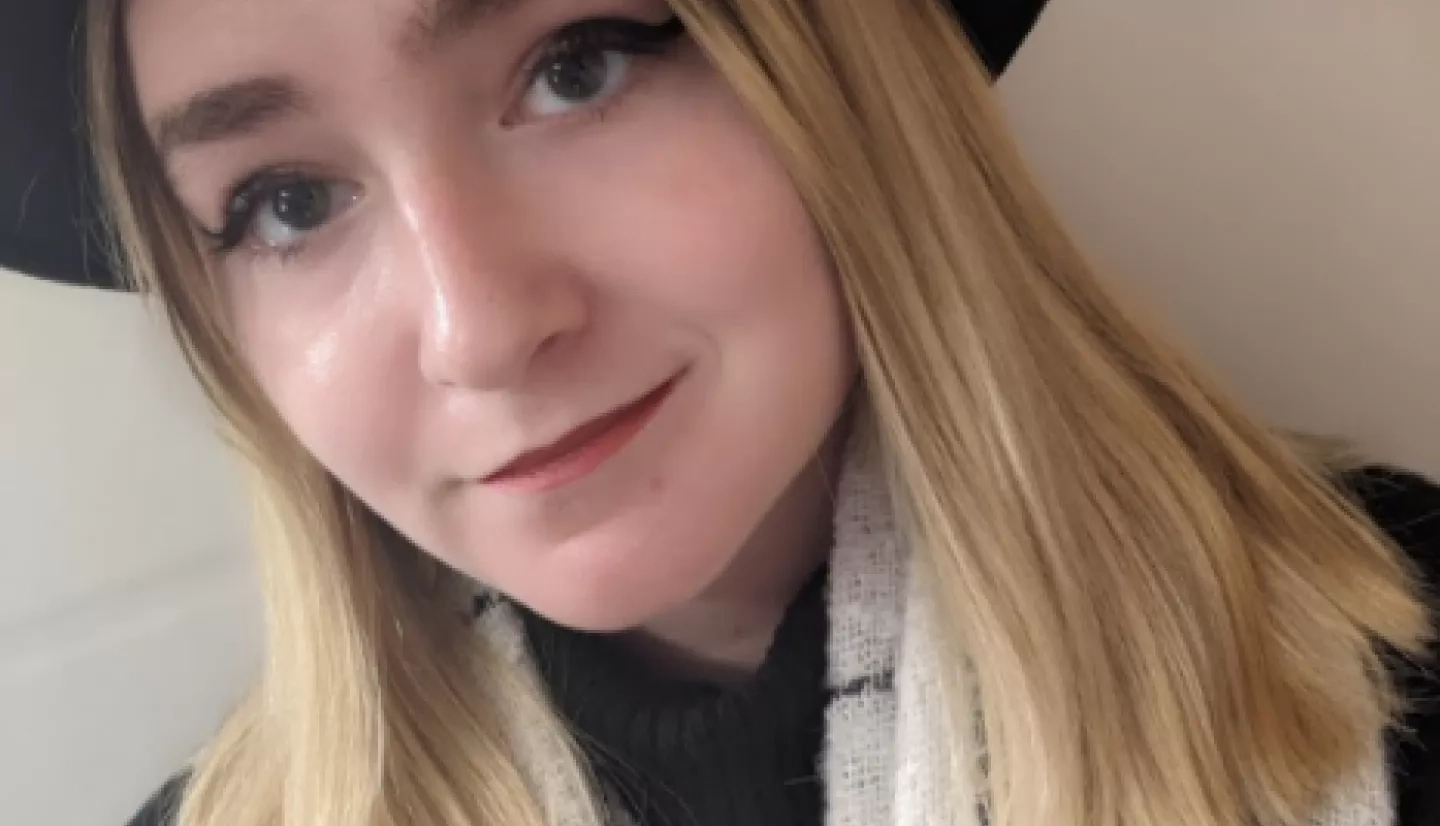 Hannah Wetzel, wearing a black hat, black and white plaid scarf, and blonde hair, smiles at the camera.