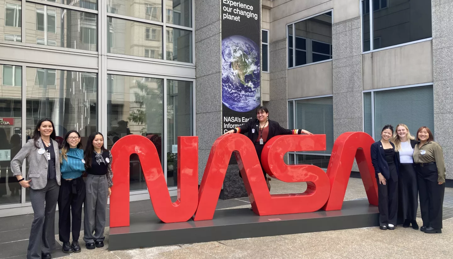 Seven professionally-dressed interns gather around the NASA logo at NASA Headquarters in Washington, DC.