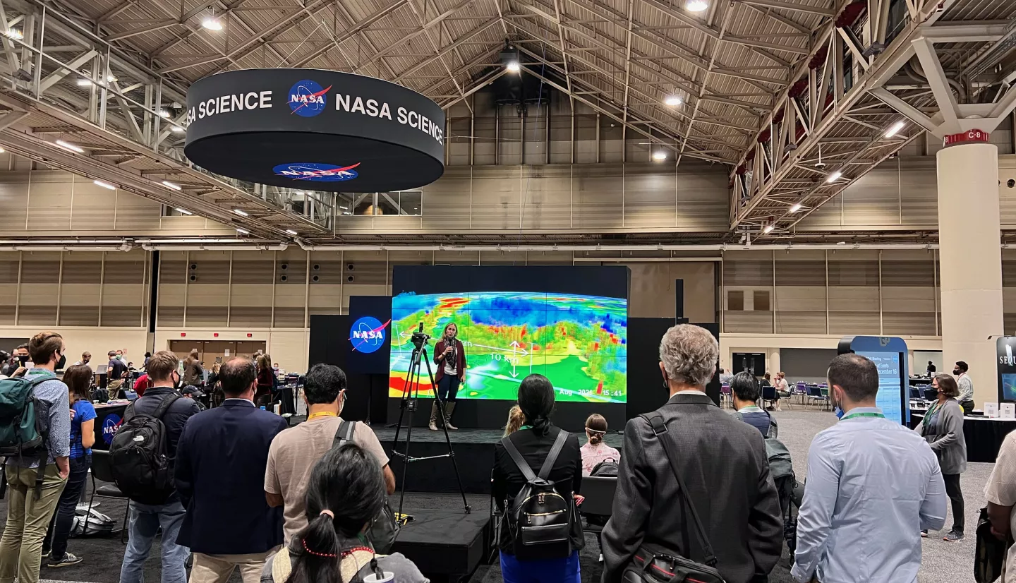 A woman named Dalia Kirschbaum stands on the stage in front of a large screen displaying data visualizations, surrounded by people watching,at the AGU 2021 Fall Meting.