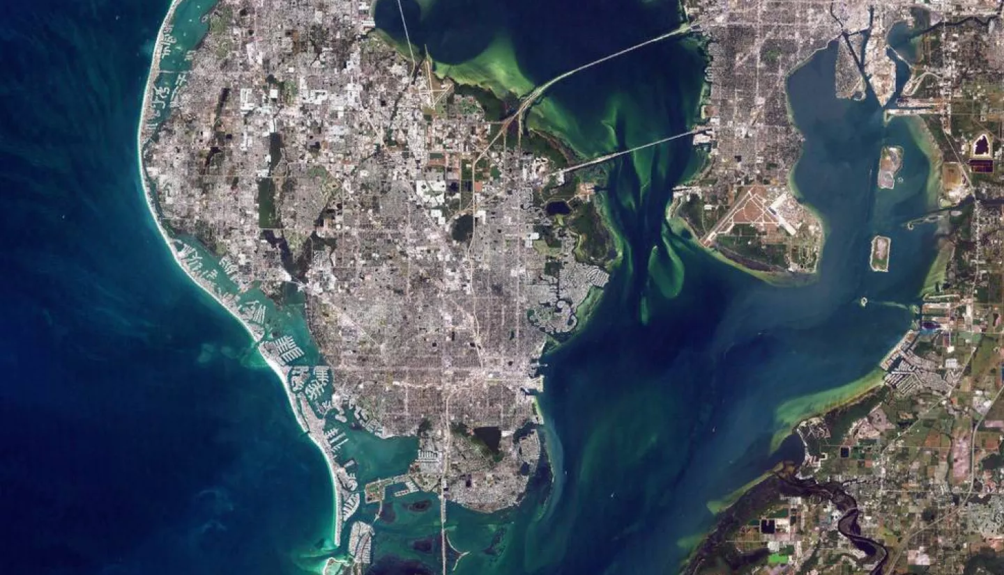 Satellite image showing the coastal city of Tampa Bay in Florida with distinct urban areas surrounded by green waters, connected by bridges over narrow channels.