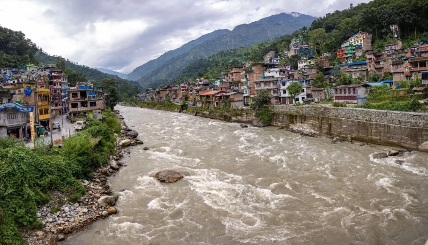 Fast-flowing river with muddy waters surges through Melamchi, Nepal, flanked by colorful houses and lush green hills under a cloudy sky.