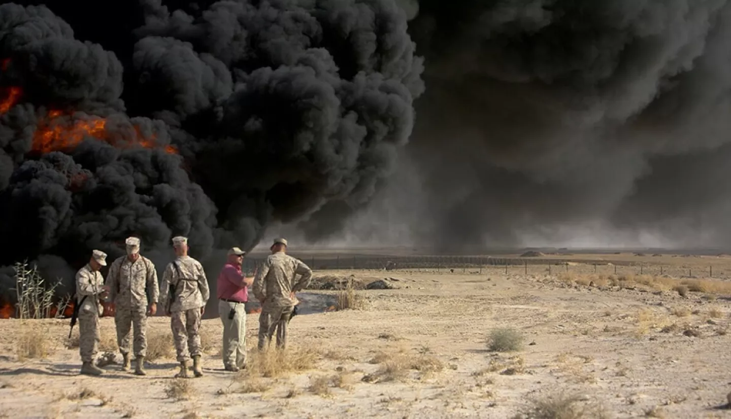 five men, four of whom are in U.S. Marine uniforms, stand in conversation near a burn pit billowing black smoke into the sky
