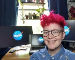 Ila White, who is wearing an astronaut-patterned button-up shirt, glasses, and curly pink hair, smiles in front of desk in their home with NASA meatball displayed on laptop and two monitors on the desk.