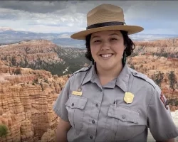 Sophie Krautmann smiles, wearing her grey National Park Ranger buttoned shirt and brown hat. She stands in a high place with the red rock formations visible in the canyon behind her.
