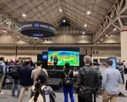 A woman named Dalia Kirschbaum stands on the stage in front of a large screen displaying data visualizations, surrounded by people watching,at the AGU 2021 Fall Meting.