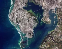 Satellite image showing the coastal city of Tampa Bay in Florida with distinct urban areas surrounded by green waters, connected by bridges over narrow channels.