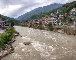 Fast-flowing river with muddy waters surges through Melamchi, Nepal, flanked by colorful houses and lush green hills under a cloudy sky.