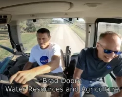 two men in the cab of a tractor with one pointing at fields as they drive along